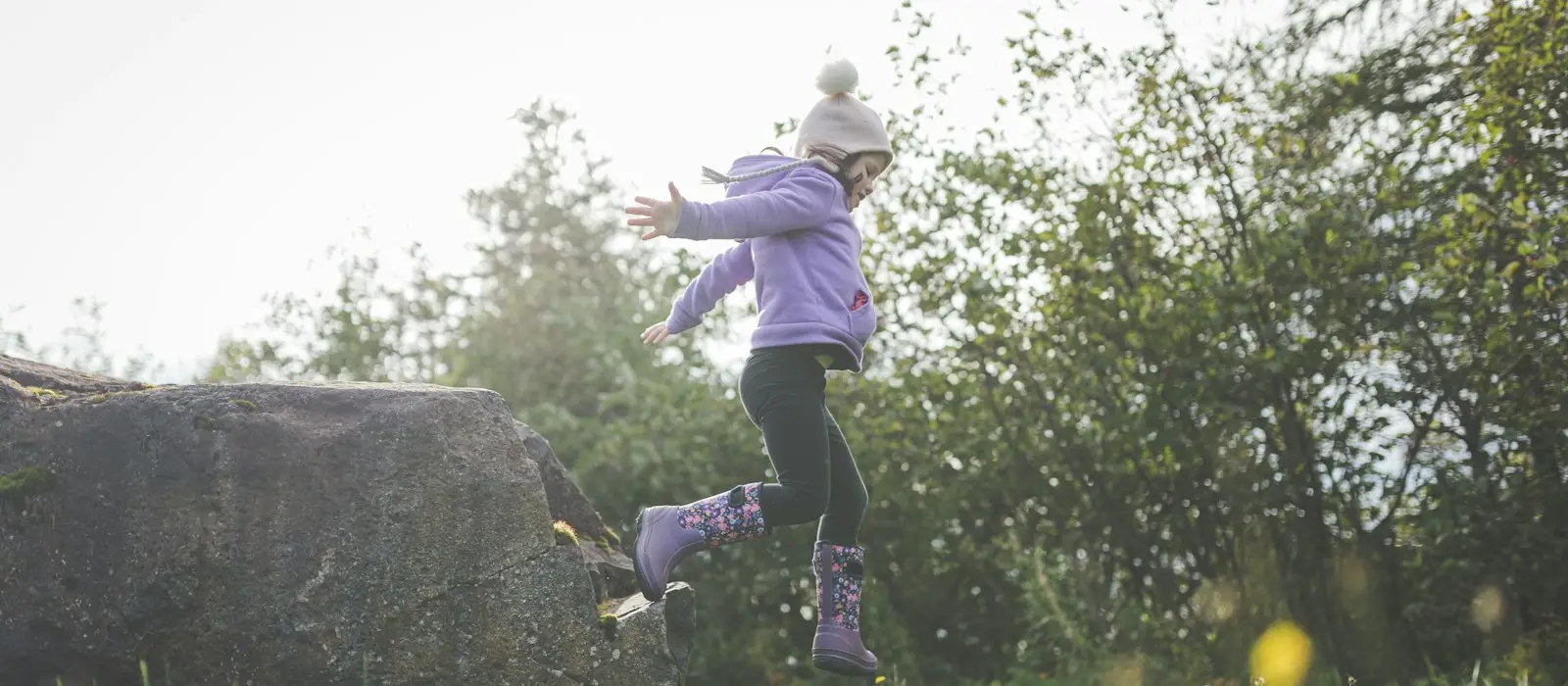 A young girl in a purple coat leaping off a rock outdoors