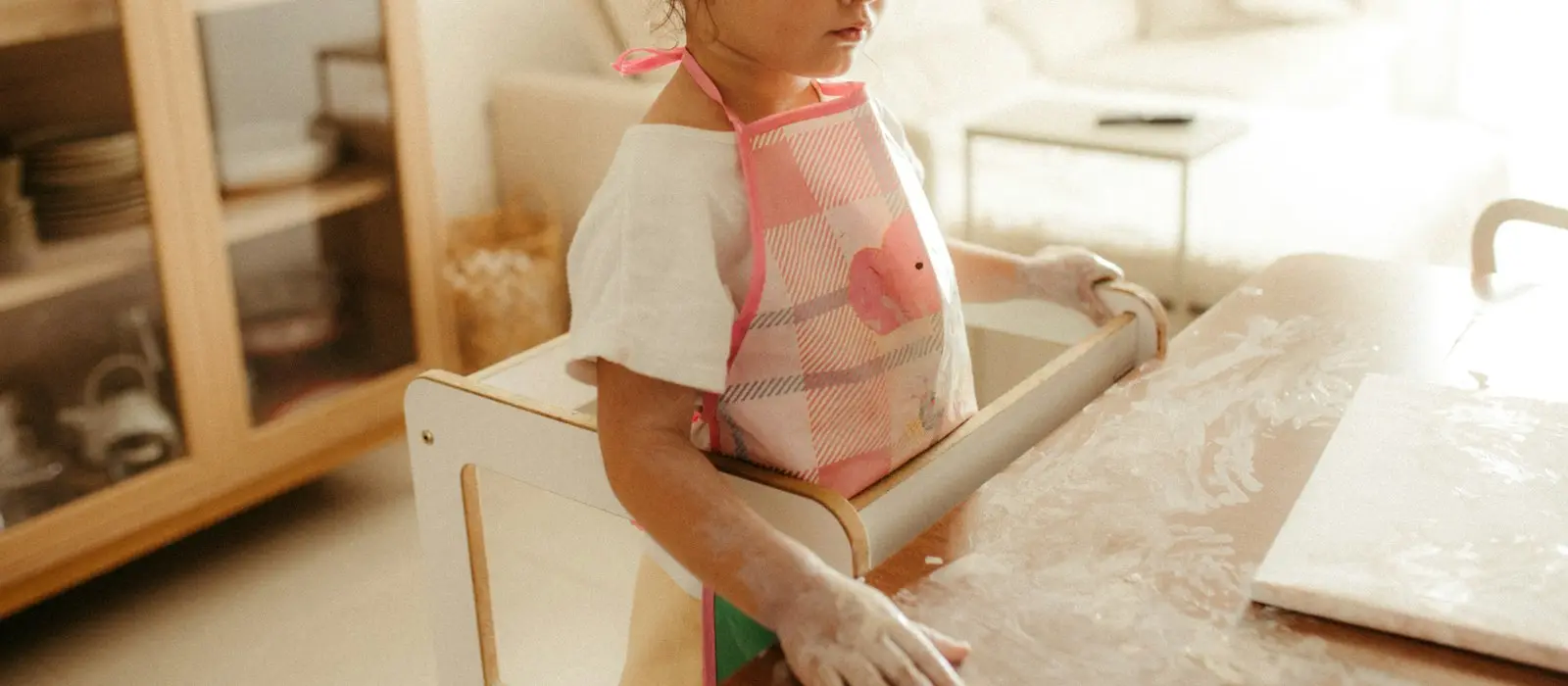 A young girl in an apron standing on a wooden learning tower, rolling out dough on a floured kitchen counter