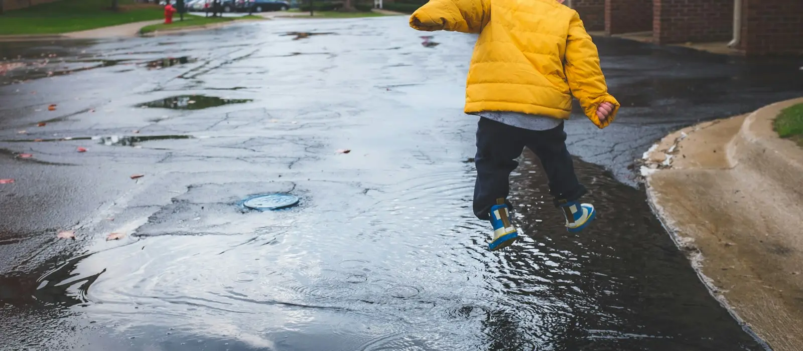 A child in a yellow raincoat jumping into a puddle on a wet street
